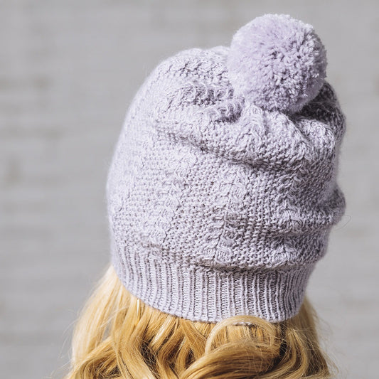 Back view close up of blonde woman wearing Delia, a pale gray cabled moss stitch hat with a pom pom in Andorra