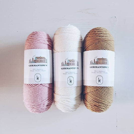 Three skeins of Kelbourne Woolens Germantown in natural, honey, and baby pink on a white wood background