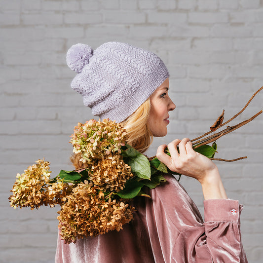 Back view of blonde woman wearing Delia, a pale gray cabled moss stitch hat with a pom pom in Andorra