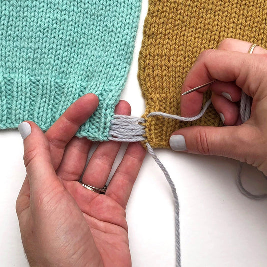 Two hands holding a darning needle and pebble Germantown seaming jade and honey stockinette stitch swatches together.