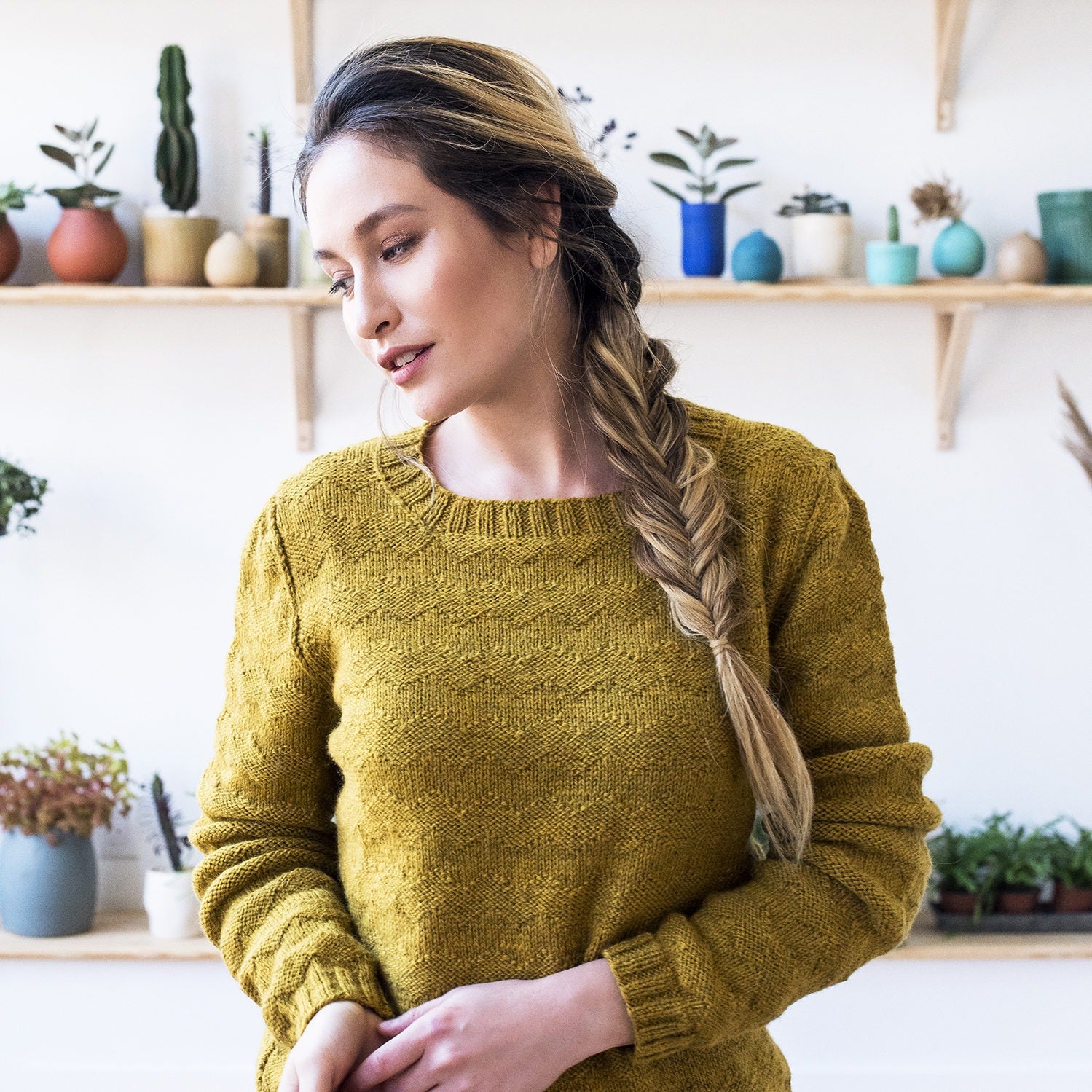 Close up of a Chinese American woman standing in a modern plant store with a long braid wearing Powell, a textured crew neck handknit sweater in Kelbourne Woolens scout in sunflower yellow.