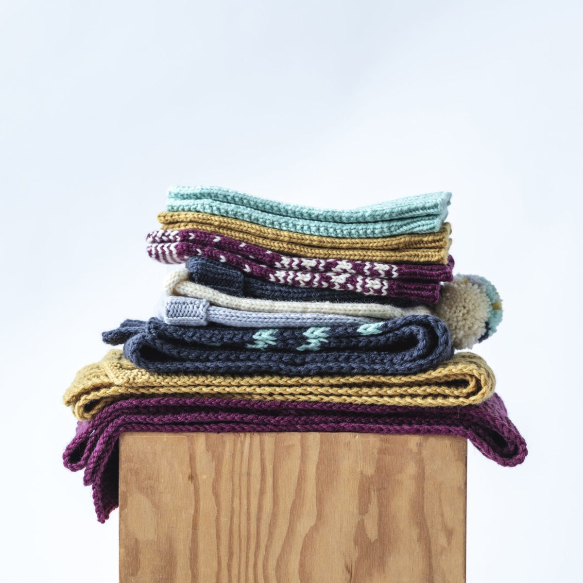 Stack of hand knit scarves, hats, and mittens in Kelbourne Woolens Germantown in Persian Red, Honey, Navy, Jade, Pebble, and Natural on a wooden box with a light blue background