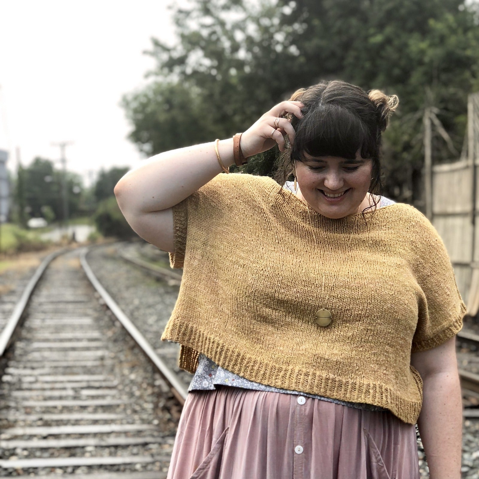Woman with brown hair wearing a gold Rift sweater by Jacqueline Cieslak standing outside near railroad tracks